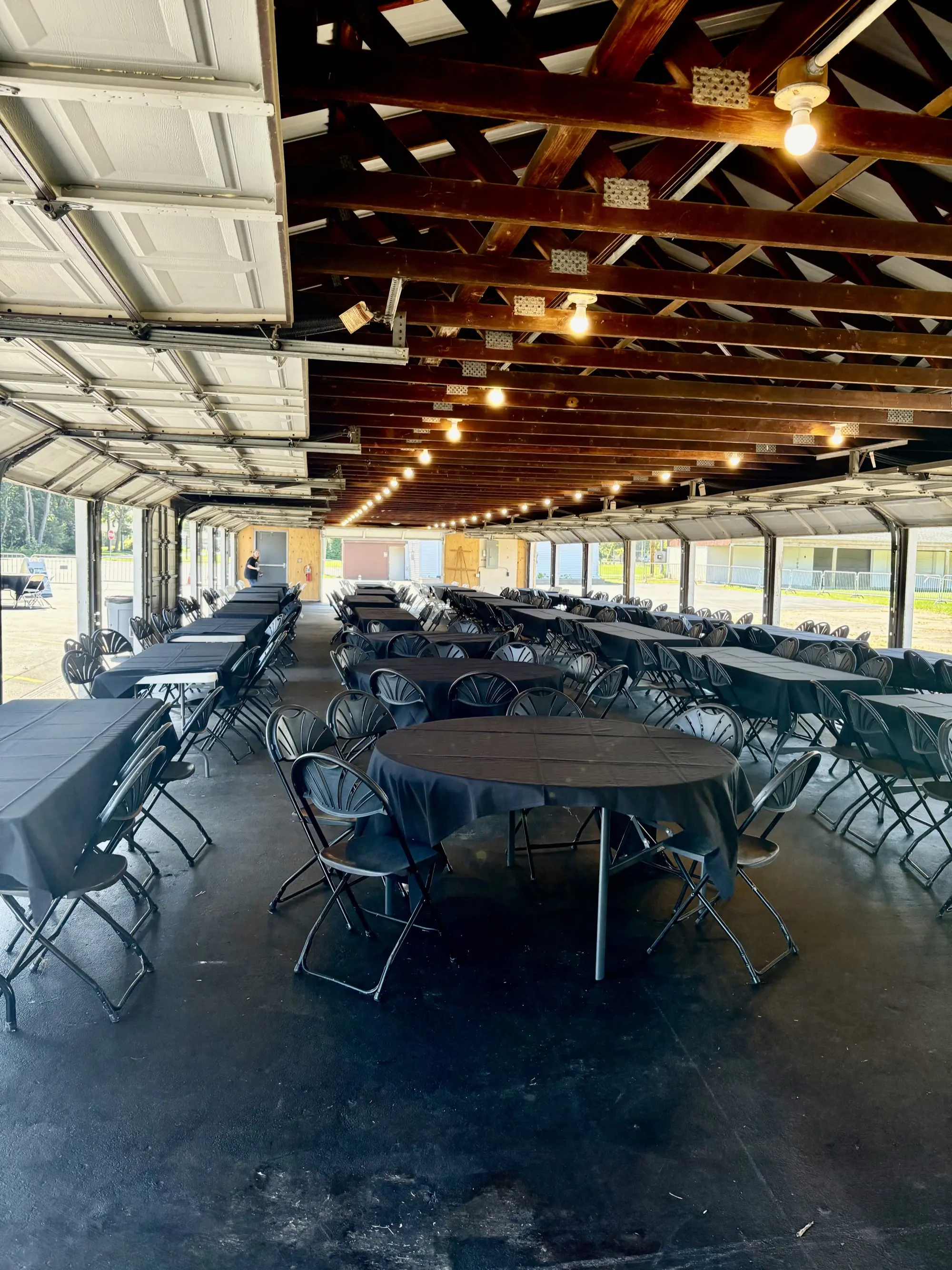 Open-air pavilion with exposed wood beam ceiling, round tables with black tablecloths, and string lights